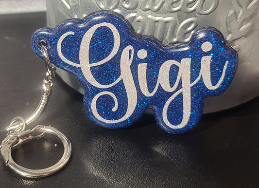 A blue handmade wooden laser-cut keychain with the word 'Gigi' laser-engraved on it, displayed against a textured background.